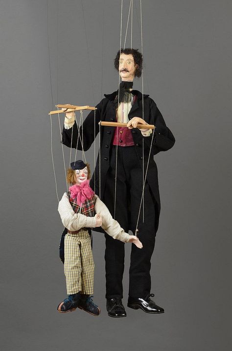 A large marionette controlling a smaller marionette. The large one wears a suit of tails. The smaller wears a clown's attire.