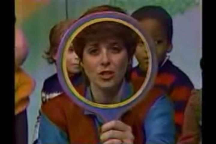 Woman looking through the frame of a purple and yellow mirror with children behind her. The image is from a 1980s children's program called Romper Room.